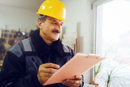 Portrait of caucasian senior man construction worker general laborer building contractor wearing yellow protective helmet holding pen and document checking data report project in the office warehouseの写真素材