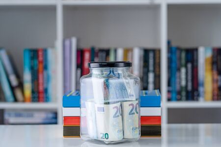 Glass jar full of 20 euro â‚¬ banknotes money and books on the white table in front of the shelf...