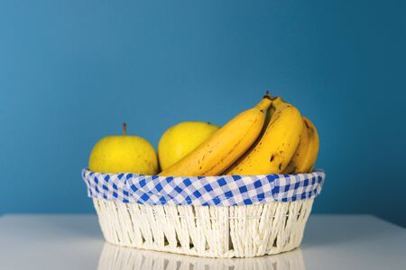 Ripe fruit bananas and apples in white basket on the table in front of the blue wall background in day organic food vegan or vegetarianの写真素材