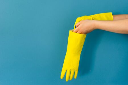 Close up on hand of unknown caucasian woman female girl putting on protective yellow rubber gloves to clean or disinfection against blue wall background in dayの写真素材