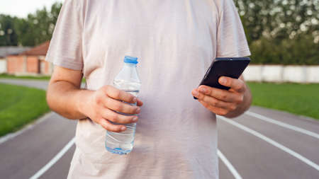 Close up on midsection of unknown caucasian man holding mobile smart phone and bottle of water on the running track - Male using application to for training progress planning and tracking in dayの写真素材