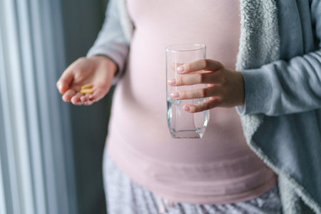Close up on midsection of unknown caucasian woman pregnant mother holding medicine supplement drugs and glass of water - taking vitamins during pregnancy conceptの写真素材