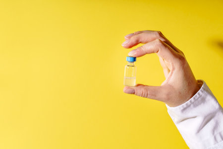 Close up on hand of unknown caucasian woman doctor holding vaccine injection in front of yellow wallの写真素材