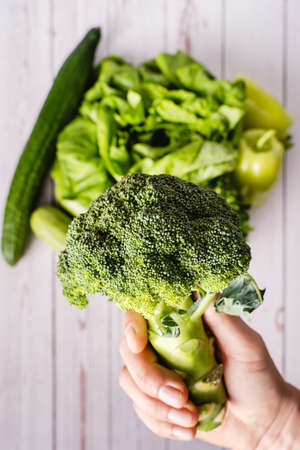 Close up on hand of unknown caucasian man holding green broccoli above table with various vegetables - bright filter raw vegan and vegetarian food healthy eating conceptの写真素材