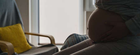 Side view on midsection of pregnant caucasian woman sitting on the bed with bed holding belly in shadow against the window - maternity pregnancy conceptの写真素材