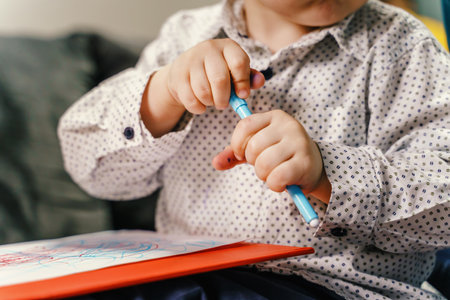 Close up on midsection of small caucasian child boy or girl hodling blue felt pen on the sofa bed at home trying to open or close it while holding in hands - childhood learning development conceptの写真素材