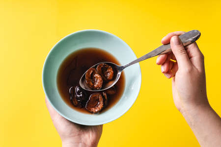 Top view on hands of unknown caucasian woman holding bowl with cooked dry plum compote over the yellow background - healthy organic food minimalistic modern abstract concept with copy spaceの写真素材