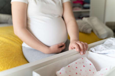 Close up on hands of unknown caucasian woman holding and packing baby clothes at home - Pregnant female preparing jumpsuit for baby in bright room - maternity motherhood and pregnancy conceptの写真素材