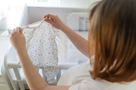 Back view on unknown caucasian woman holding a baby clothes by the cradle at home - Pregnant female preparing jumpsuit for baby in bright room - maternity motherhood and pregnancy conceptの写真素材