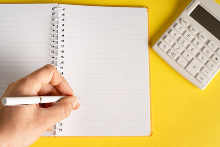 close up on hand holding pencil writing on notebook by the calculator on yellow background - office and business or goals conceptの写真素材