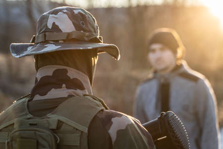Back view of unknown armed soldier standing against a migrant or terrorist or criminal in sunny...