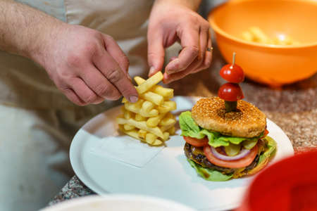 Close up on hands of unknown caucasian man chef making burger stacking potato french fries chips on the counter-top in the kitchen at restaurant or at homeの写真素材
