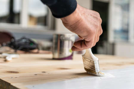 Close up on white paint brush in the hand of unknown man painting wooden board hobby and renovation repairing conceptの写真素材