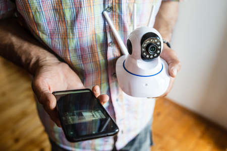 Close up on midsection of unknown man holding home security surveillance camera and mobile phone...