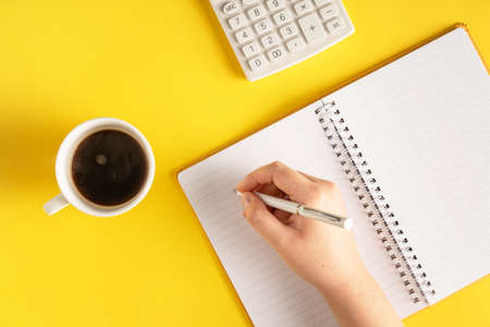 close up on hand holding pencil writing on notebook by the calculator on yellow background - office...