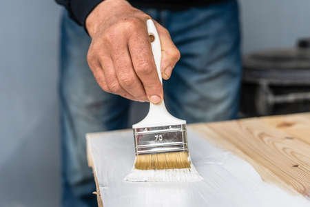 Close up on white paint brush in the hand of unknown man painting wooden board hobby and renovation repairing conceptの写真素材