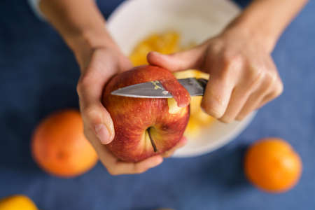 Close up on hands of unknown caucasian woman peeling red apple over the bowl on the table at home - healthy eating concept organic food homemade vegetarian or vegan meal - top viewの写真素材