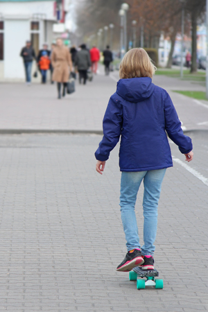 Girl riding a skateboard on a city street. A child of a pre-teen age on a walk. Girl dressed in a jacket and jeans. View from the back.の写真素材