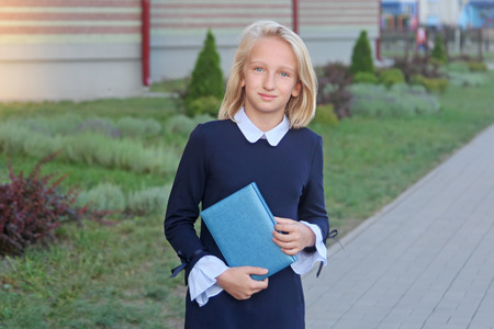 Beautiful blonde girl in elegant dress with book in hands near school. Schoolgirl likes to learn and...
