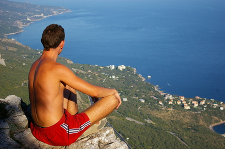 Young man sitting on a rock and looking down on the sea coastの写真素材