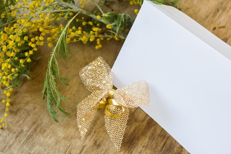 Beautiful yellow mimosa and a card with a gold bow on a stylish wooden backgroundの写真素材