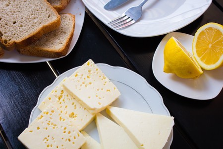 various pieces of cheese lying on a plate next to the lemon and slices of brown bread on a white plate and a stylish black table.の写真素材