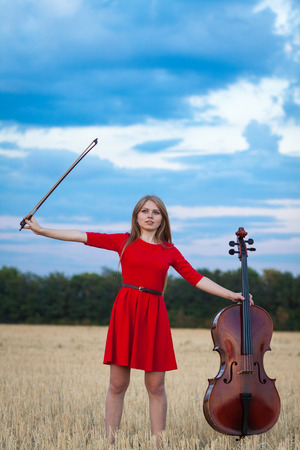 Beautiful woman professional musician in red dress with cello outdoorsの写真素材