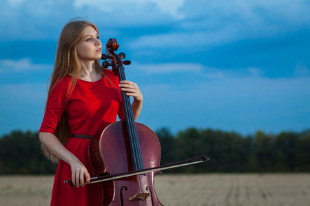 Beautiful woman professional musician in red dress with cello outdoorsの写真素材