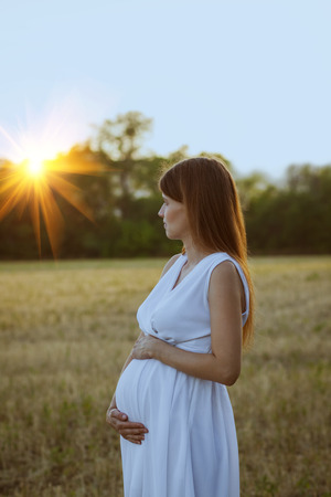 Pregnant woman in white dress watching the sunset outdoorsの写真素材