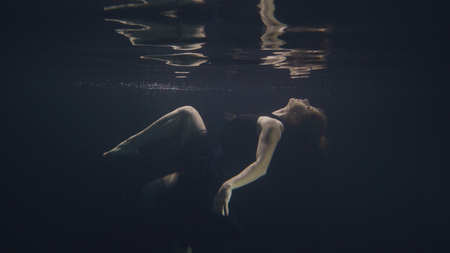 Young woman in black dress swimming underwater in pool while video shootingの写真素材