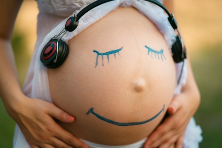 Pregnant woman belly closeup with smiling funny face drawing on it, listening to the music in earphonesの写真素材