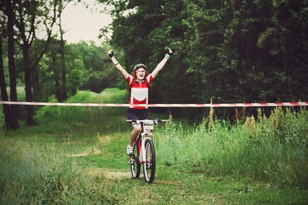 Young red-haired woman cyclist in protective helmet crossing the finish line breaking the tape.の写真素材
