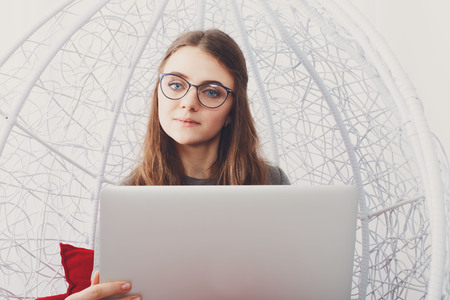 Young beautiful woman in eyeglasses in fashionable armchair with laptop. Girl with pc in modern interior at red cushions.の写真素材