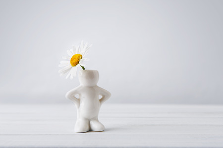 Beautiful daisy in small white ceramic vase in form of man on white table at neutral background. Small man with flower head. Flower with copy space. Funny vase.の写真素材