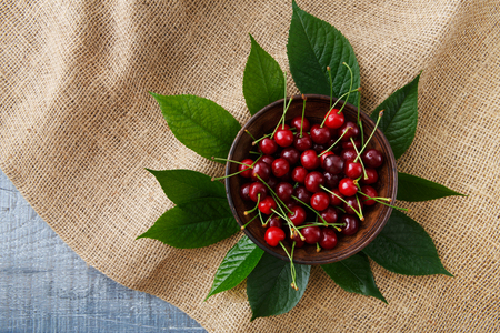 Sweet fresh cherries in a bowl with green leaves on blue rustic wood and sack cloth, fruit backround. Healthy food top view at hessian textile.の写真素材