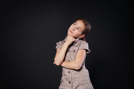 Thinking child portrait. Little preteen girl pensive, consider something, looking up. Primary school student solving some problem at black studio background with copy space.の写真素材