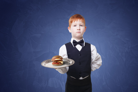 Little waiter stands with tray serving hamburger. Redhead child boy in suit plays restaurant servant, gives burger at blue backgroundの写真素材