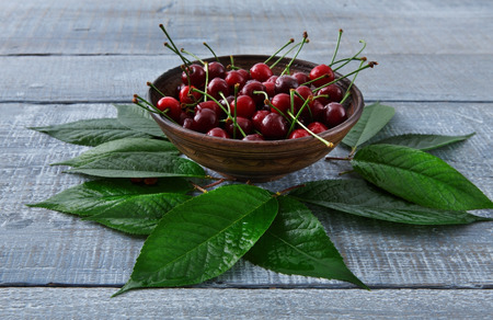 Sweet fresh cherries in bowl with green leaves background. Scattered fruits on blue rustic wood table. Healthy food.の写真素材