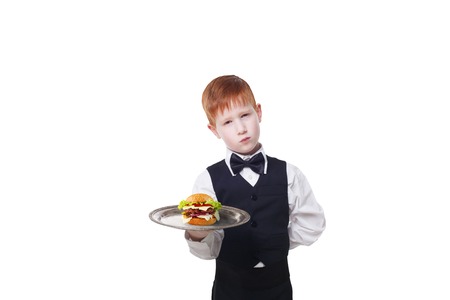 Little sad and tire waiter stands with tray serving hamburger. Redhead child boy in suit plays hardworking restaurant servant, gives burger isolated at white backgroundの写真素材