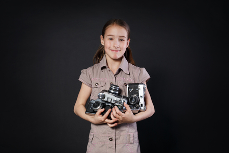 Little cute girl pose with heap of old vintage film cameras at black background. Small child photographer happy and smiling, studio portrait. Collecting antiques, old thingsの写真素材
