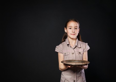 Little smiling caucasian girl holds empty stainless food tray. Female child offering somehting, studio portrait at black background.の写真素材