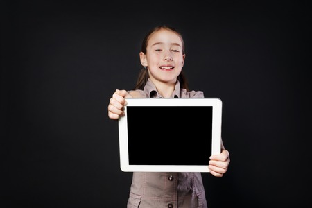 Little happy girl shows empty screen of white Digital Tablet, place for copy space. Female child studio portrait wih modern device at black backgroundの写真素材