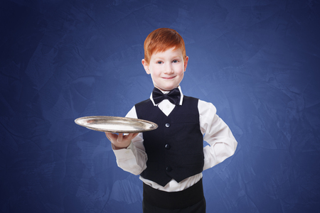 Little waiter stands with empty tray serving. Smiling redhead child boy in suit plays restaurant...