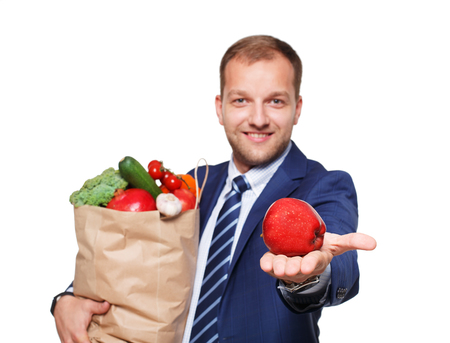Smiling young businessman hold shopping bag full of groceries isolated at white background. Healthy food shopping. Paper package with vegetables and fruits, happy man buyer show red appleの写真素材