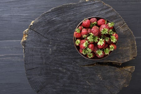 Red fresh strawberries on black rustic wood background. Bowl with natural ripe organic berries with peduncles on wooden circle cut out, top view with copy spaceの写真素材