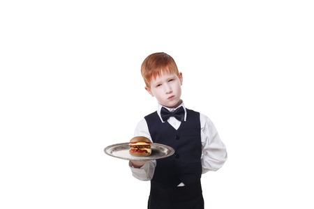 Little sad and tire waiter stands with tray serving hamburger. Redhead child boy in suit plays hardworking restaurant servant, gives burger isolated at white backgroundの写真素材