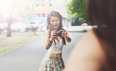 Young asian girl taking photo of her friend outdoors with digital camera. Young female tourist in boho fashion clothes, laughing and having fun in summer park. Travelling together, lifestyle portrait.の写真素材