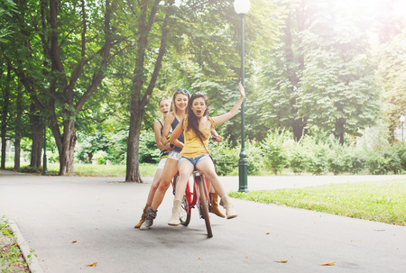 Happy boho chic stylish girls ride together having fun. Beautiful women on bicycles with basket full of wild flower. Female friends, youth and happiness, active summer leisure in park concept.の写真素材