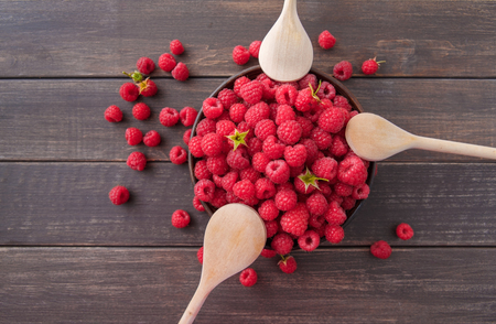 Red fresh raspberries on brown rustic wood background. Bowl with natural ripe organic berries with peduncles and wooden spoons on table, top view, flat lay with copy spaceの写真素材