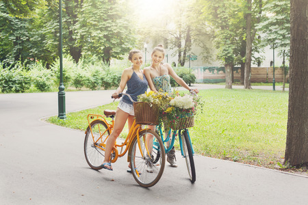 Happy boho chic stylish girls ride together having fun. Beautiful women on bicycles with basket full of wild flower. Female friends, youth and happiness, active summer leisure in park concept.の写真素材